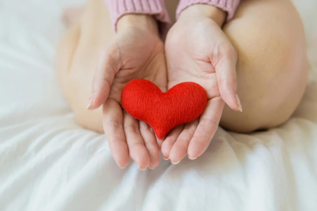 Hands gently cupping a small red heart-shaped plush on a white bed, with a person in a pink sweater sitting cross‑legged in the background.